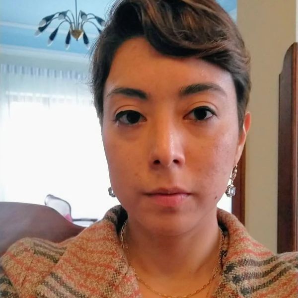 Close-up portrait of a woman with a brown pixie-cut and dark brown eyes, seated indoors, facing the camera.