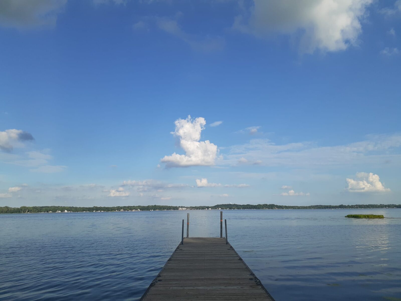 Image of a a blue lake with a dock jutting out in the center. Sky is blue with a few white clouds. 