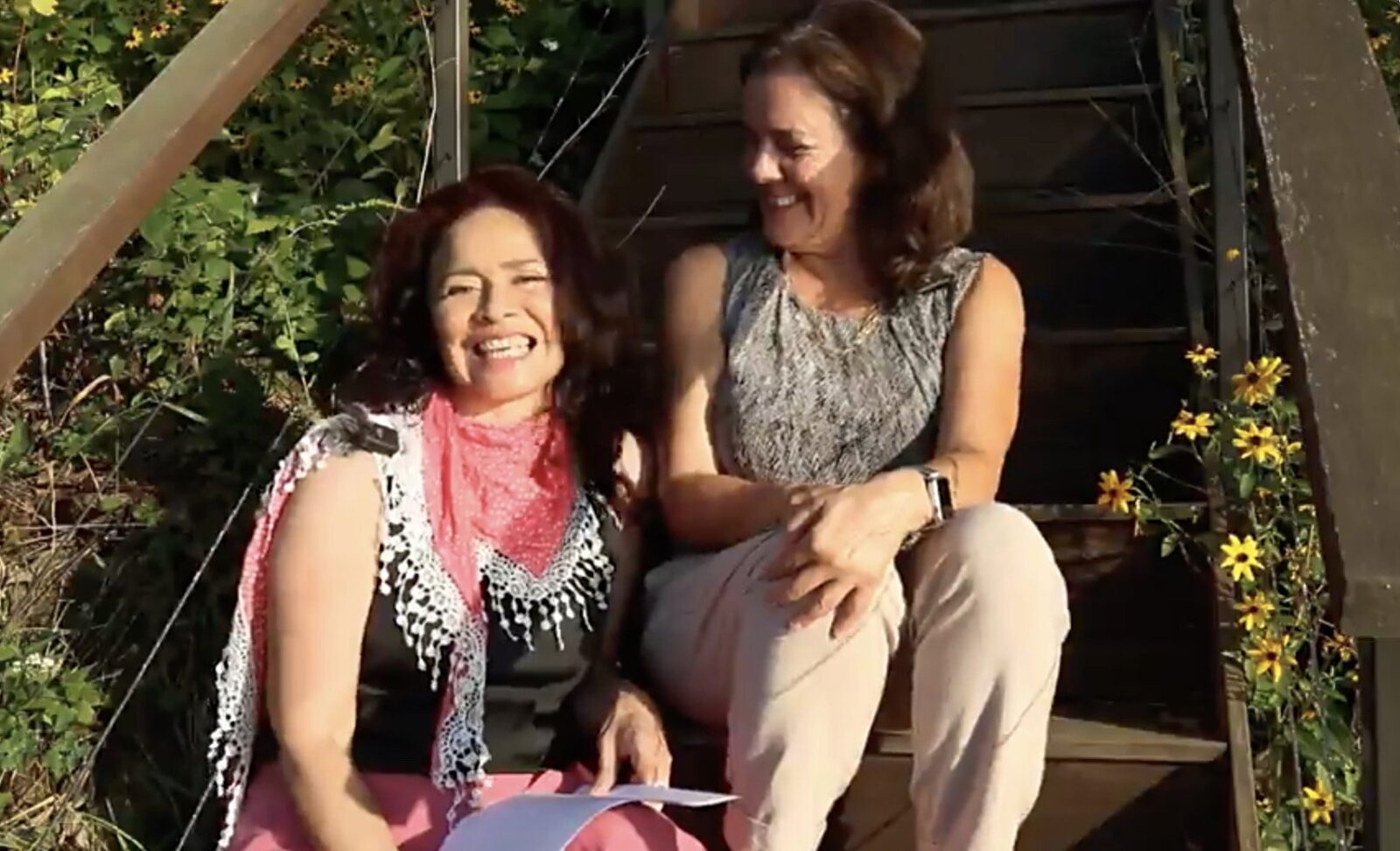 Sarlie E. Mercado (left) and Lori DiPrete Brown (right) sit on a wooden outdoor staircase framed by green foliage and small yellow flowers. One Sarlie holds a sheet of paper while both are positioned near the center of the steps in bright sunlight
